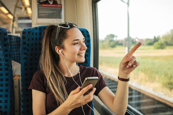 Junge Frau in der Bahn mit Handy und Kopfhörern deutet auf Fenster