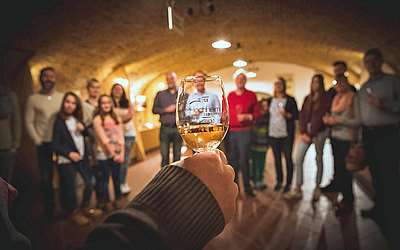 Vergrößerte Ansicht: Hand hält ein gefülltes Weinglas, dahinter eine Besuchergruppe in einem Gewölbe-Keller