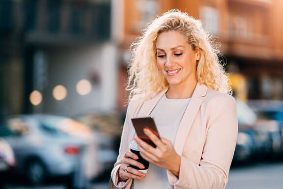 lächelnde Frau auf der Straße mit Smartphone und Coffe-to-go-Becher in der Hand