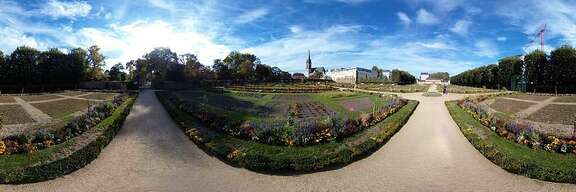 Panoramabild: 3 angelegte Beetfelder, dazwischen Wege aus Kieselsand. Im Hintergrund Gebäude und sehr blauer Himmel mit Schleierwolken