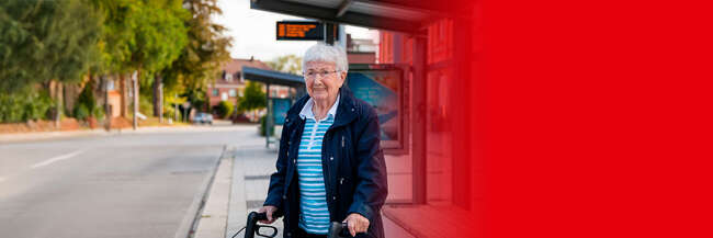 Teaserbild Rollator-Training Eine ältere Frau mit kurzem weißen Haar und Brille steht an einer Bushaltestelle, hält einen Rollator und lächelt in die Kamera.