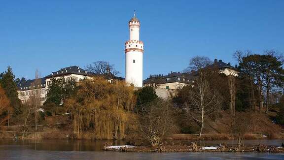 Sicht auf einen See mit kahlen Bäumen, im Hintergrund ragt ein weißer historischer Turm in die Höhe.