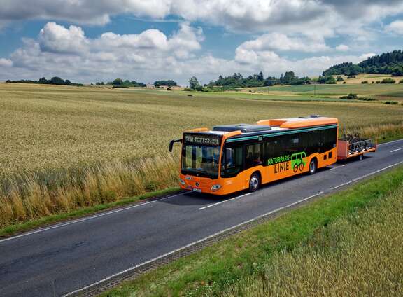 Bus in oranger Farbe mit Fahrradanhänger abgebildet in einer ländlichen Umgebung