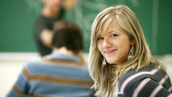 A girl in a classroom looking in the camera, behind her a teacher showing things on a blackboard