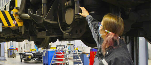 Eine Frau zeigt etwas an einem Eisenbahnrad eines hochgebockten Zuges 