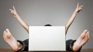 A man sits hidden behind a laptop. You can only see his arms and feet. 
