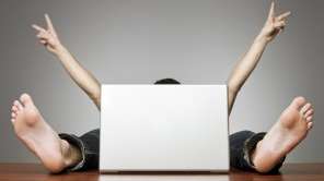 A man sits hidden behind a laptop. You can only see his arms and feet.