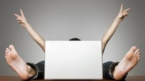 A man sits hidden behind a laptop. You can only see his arms and feet.