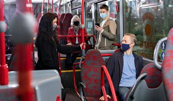 Menschen mit Mund-Nasen-Bedeckung stehen und sitzen im Innenraum eines Busses mit roten Sitzen