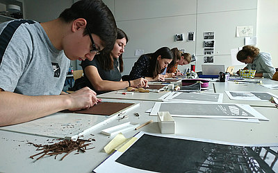 Vergrößerte Ansicht: Mehrere Schüler gestalten an einem langen Tisch sitzend Linoldrucke