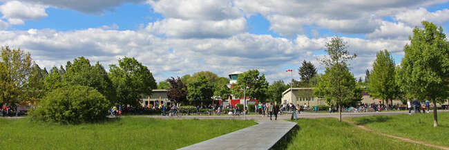 Holzsteg führt auf eine asphaltierte Straße. Im Hintergrund Gebäude und Tower.