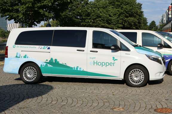 Ein weißer Van steht auf einem Platz unter Bäumen, Aufschrift: On-Demand-Shuttle, kvgOG Hopper