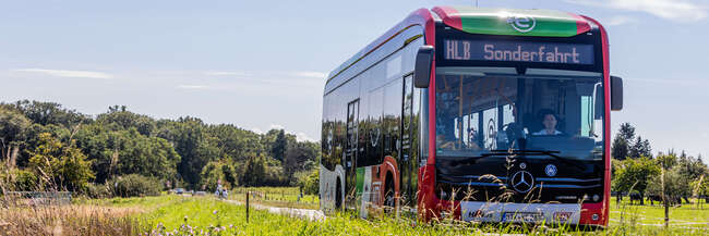 Ein Elektrobus der MTV fährt auf einer Landstraße durch eine grüne Landschaft im Main-Taunus-Kreis. Im Vordergrund sind Wiesenblumen zu sehen, im Hintergrund Bäume unter blauem Himmel.