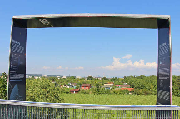 Vergrößerte Ansicht: Informationstafel in Form eines Tores mit Blick ins Grüne, im Hintergrund Häuser und Skyline