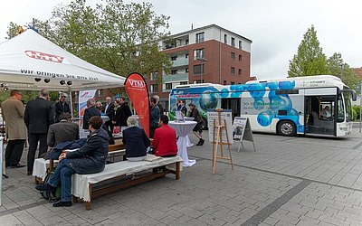 xEine Gruppe Menschen hört Herrn Baron bei einem Vortrag zu. Ein Elektrobus befindet sich im Hintergrund.