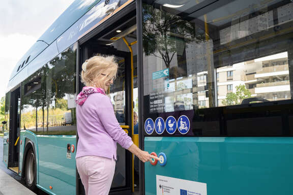Eine Seniorin steht an einem Bus und drückt eine Taste.
