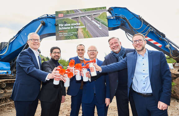 Hinten zwei blaue Bagger, vor denen ein Plakat mit der Aufschrift "Für eine starke Schiene in Hessen. Vier Gleise von Hanau nach Gelnhausen. Im Vordergrund sechs Männer mit Tröten in Hand, auf denen das Logo der Deutschen Bahn zu sehen ist.