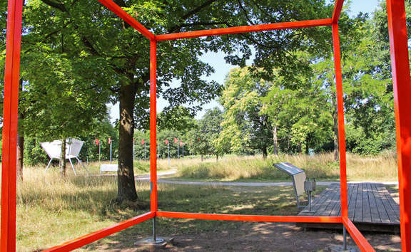 Vergrößerte Ansicht: orange Metallstangen zu einem Würfel gesteckt vor Wiese mit Bäumen