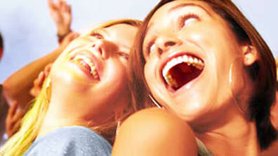 A blonde laughing woman leans her head back and next to her stands a brunette laughing woman back to back with her. They both look at each other. It seems as if they are at a party and are having a good time. In the background, there is a crowd of people celebrating as well. 