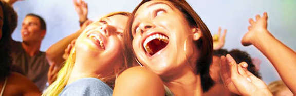 zwei junge Frauen lächeln sich fröhlich an, im Hintergrund erhobene Hände von feiernden Menschen