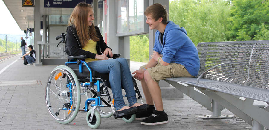 Junge Frau im Rollstuhl am Bahngleis mit einem männlichen Begleiter
