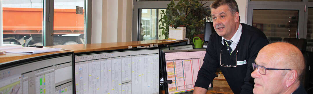 Ein Mann sitz vor vielen Monitoren am Schreibtisch, ein anderer Mann lehnt sich an den Tisch und schaut mit