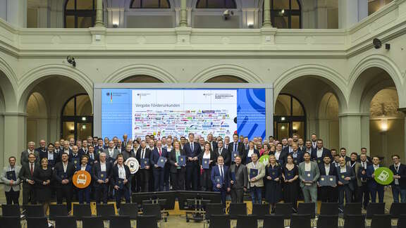 Große Gruppe von Menschen steht in einem historischen Saal auf einer Bühne und hält Förderurkunden in blauen Mappen. Im Hintergrund zeigt eine Leinwand die Überschrift „Vergabe Förderurkunden“ mit vielen Logos von Verkehrsunternehmen.
