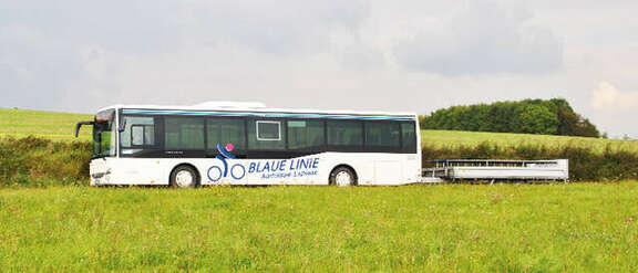 Vergrößerte Ansicht: Ein weißer Bus der Blauen Linie mit Fahrradanhänger fährt durch eine sommerliche Landschaft