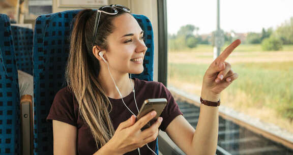 Junge Frau in der Bahn mit Handy und Kopfhörern deutet auf Fenster