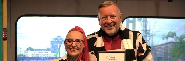 RMV-Lieblingsbusfahrerin 2025: Justyna Cielecka-Drozdz aus Hanau Ein Mann hält eine Urkunde in der Hand, eine Frau steht daneben mit einem Blumenstrauß.