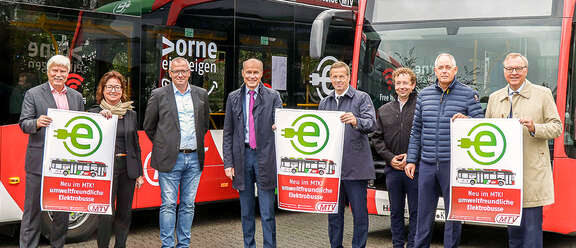 Teilnehmende des Pressetermin stehen vor den neuen Elektrobussen und halten ein Plakat hoch: "Neu im MTK! umweltfreundliche Elektrobusse"