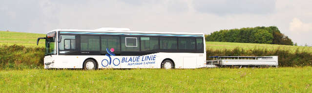 Vergrößerte Ansicht: Ein weißer Bus der Blauen Linie mit Fahrradanhänger fährt durch eine sommerliche Landschaft
