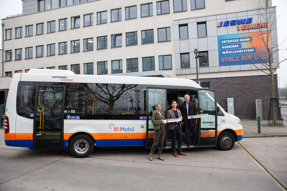 Ein Shuttle-Bus, in dem ein Mann steht. Vor dem Bus stehen zwei weitere Personen, die ein Plakat in den Händen halten
