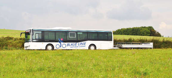 Vergrößerte Ansicht: Ein weißer Bus der Blauen Linie mit Fahrradanhänger fährt durch eine sommerliche Landschaft