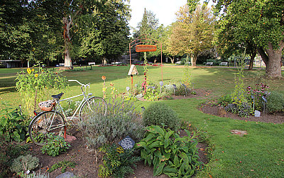 Vergrößerte Ansicht: Beete mit Kräutern und Blumen auf Wiese, umgeben von Bäumen. Altes Fahrrad im vorderen Beet