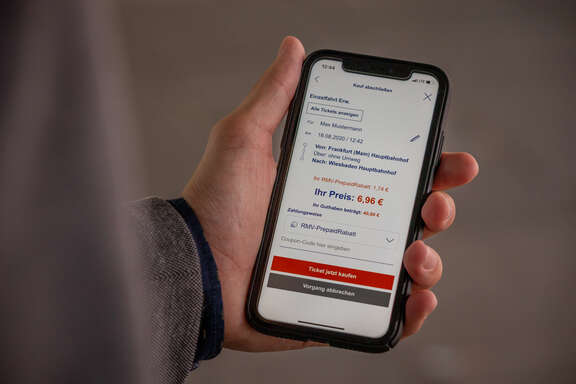 RMV-PrepaidRabatt Eine Hand hält ein Handy, im Display ist ein Fahrkartenkauf mit PrepaidRabatt zu sehen