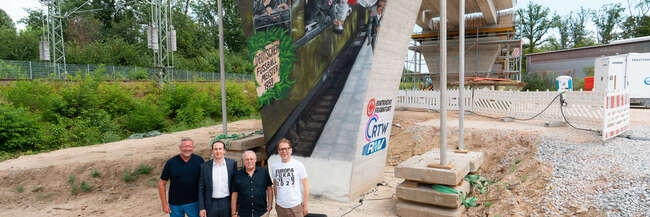 Vor dem Brückenpfeiler (v.l.n.r.): Prof. Knut Ringat (RMV), Julien Zamberk (Eintracht Frankfurt), Horst Amann (RTW Planungsgesellschaft) und der Frankfurter Mobilitätsdezernent Wolfgang Siefert (v. l.) vor dem von Fan-Vertretern der Eintracht Frankfurt gestalteten Mural am Frankfurter Waldstadion.