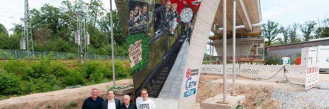 Vor dem Brückenpfeiler (v.l.n.r.): Prof. Knut Ringat (RMV), Julien Zamberk (Eintracht Frankfurt), Horst Amann (RTW Planungsgesellschaft) und der Frankfurter Mobilitätsdezernent Wolfgang Siefert (v. l.) vor dem von Fan-Vertretern der Eintracht Frankfurt gestalteten Mural am Frankfurter Waldstadion.