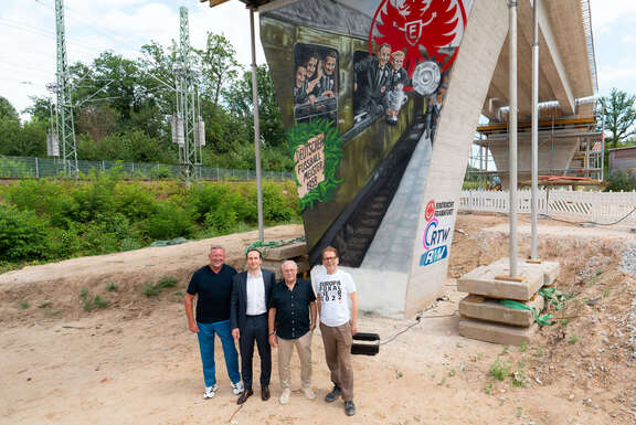 Vor dem Brückenpfeiler (v.l.n.r.): Prof. Knut Ringat (RMV), Julien Zamberk (Eintracht Frankfurt), Horst Amann (RTW Planungsgesellschaft) und der Frankfurter Mobilitätsdezernent Wolfgang Siefert (v. l.) vor dem von Fan-Vertretern der Eintracht Frankfurt gestalteten Mural am Frankfurter Waldstadion.