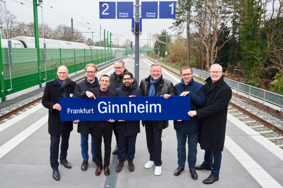 Acht Männer stehen nebeneinander stehen in einer S-Bahn-Station und halten ein blaues Schuld mit der Aufschrift Ginnheim.