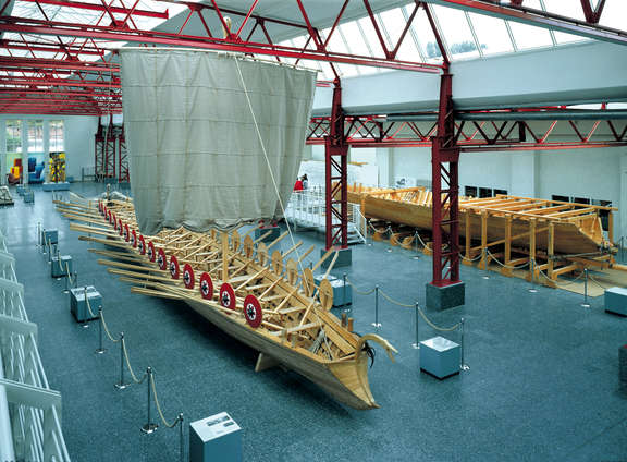 Museum für Antike Schifffahrt Mainz In einer großen Halle mit roten Trägern steht ein gewaltiges antikes Schiff mit Segel und Schutzschildern