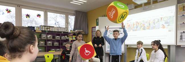 Kinder in einer Schulklasse sitzen in einem Stuhlkreis und spielen mit zwei Würfeln, auf denen ÖPNV-Zeichen zu sehen sind.