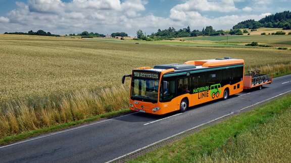 Bus in oranger Farbe mit Fahrradanhänger abgebildet in einer ländlichen Umgebung