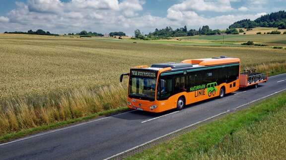 Bus in oranger Farbe mit Fahrradanhänger abgebildet in einer ländlichen Umgebung