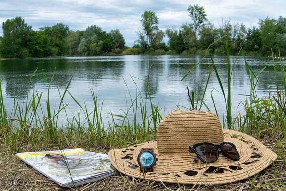 Strohhut, Sonnenbrille, Armbanduhr und Zeitschrift im Gras vor Badesee