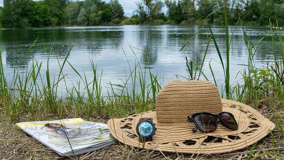 Strohhut, Sonnenbrille, Armbanduhr und Zeitschrift im Gras vor Badesee