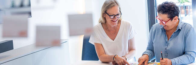 Zwei Mitarbeiterinnen sitzen gemeinsam an einem Tisch und besprechen Unterlagen. Beide lächeln und zeigen auf bunte Informationsmaterialien, die vor ihnen liegen.