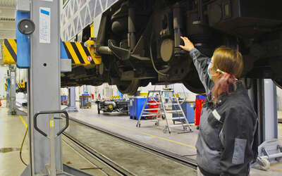 Eine Frau zeigt etwas an einem Eisenbahnrad eines hochgebockten Zuges 
