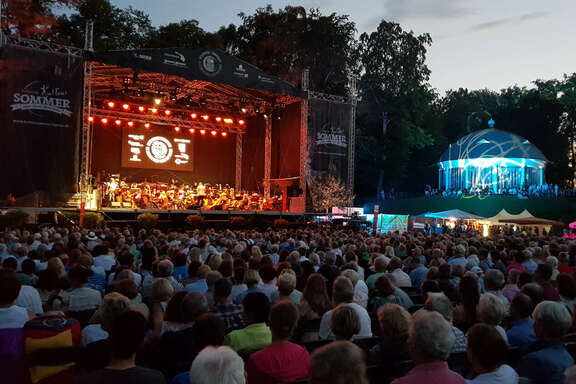 Open-Air-Bühne vor Bäumen und Publikum in der Abenddämmerung
