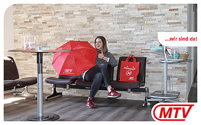 Eine Frau mit rotem MTV-Regenschirm sitzt auf einem Stuhl im Empfangsbereich: ...wir sind da!